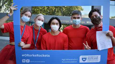 Group of 5 students in red shirts stand behind a blue selfie frame in front of the city campus library