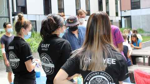 Three RAs are helping a group of residents with the BBQ. They have their back to the camera so we can see how pronouns have and have not been printed on their staff shirts. 