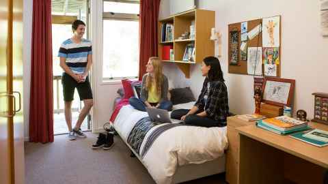 Three students in a room with two sitting on the bed and one standing in the doorway