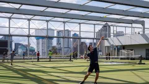 One person in the foreground prepares to serve a tennis ball on a green turn inside Hiwa. Other people appear in the distance