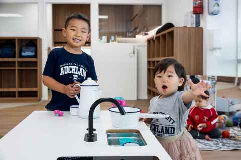 Two children next to a play kitchen sink smiling towards the camera. Both are wearing University of Auckland shirts.