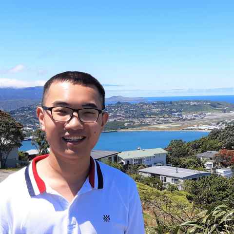 Male student in white shirt and red, blue and white collar smiling for a photo with Rangitoto Island seen in the background