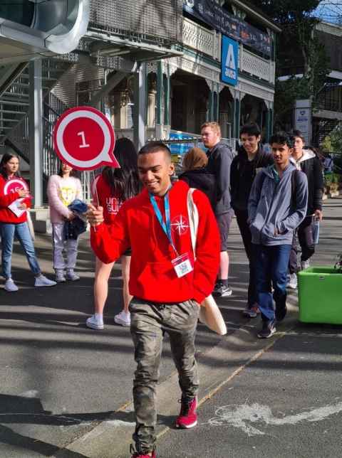 Male student in red UniGuide sweatshirt holding a paddle with the number '1' on it leading a group of students outside AUSA House at City Campus. Another small group of students can be seen in the background.