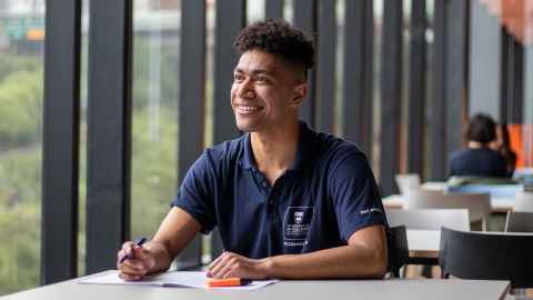 Person sits a table in a blue shirt with University Accommodation logo looking off in the distance and smiling