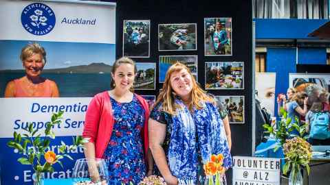 Two people at an expo promoting their volunteer work stand behind their table and smile at the camera
