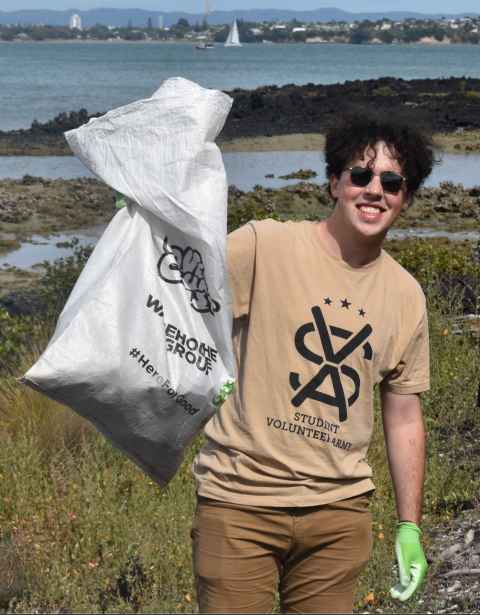 Person stand in front of a large body of water wearing a Student Volunteer Army t-shirt. They are holding up a big white bag and smiling. 