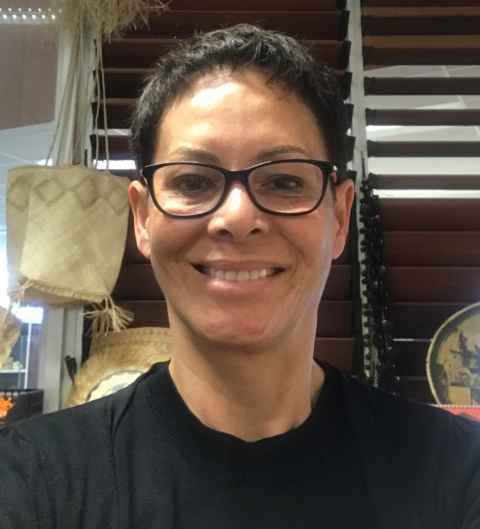 A smiling Pacific woman wearing glasses, with flax bags and other items in the background.