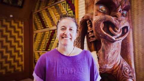 A smiling woman in a purple t-shirt, in front of a Māori carving.