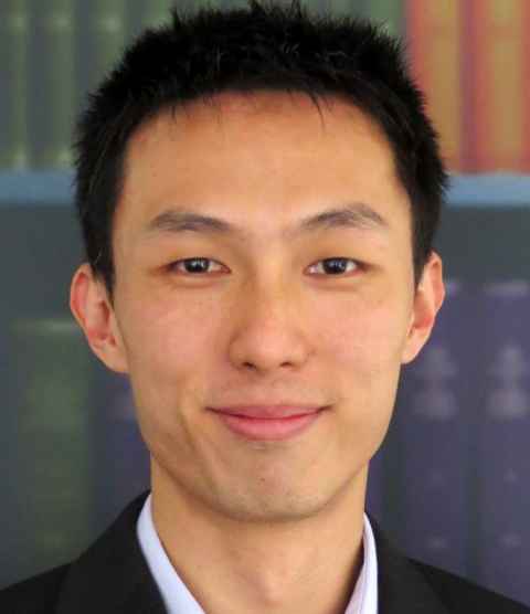 A close-up of a man in a pale shirt and black jacket, with bookshelves in the background.