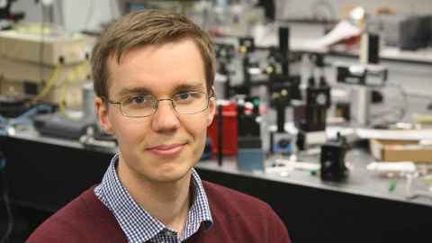 A young man wearing glasses, in a lab full of equipment.