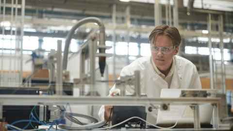 A young man in a white lab coat and protective glasses, using engineering lab equipment.