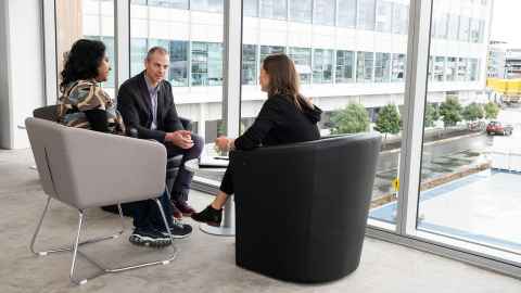 Three people seated in a circle, beside a window with a city view.