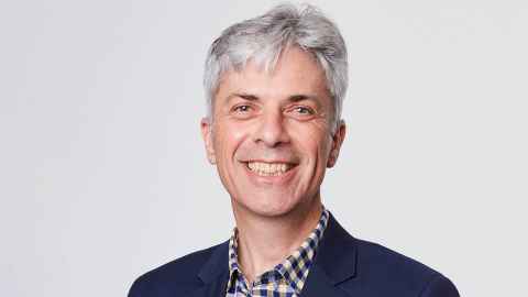 Portrait of Frank, who has a narrow face, short grey hair, longer at the fringe and is wearing a suit coat, with a checked shirt, open at the neck.