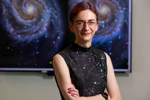 Professor Jan J. Eldridge, Head of the Department of Physics, in a black sleeveless dress with tied back hair, standing with arms crossed in front of a screen showing a picture of the universe.