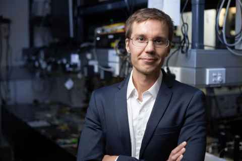 Professor Miro Erkintalo, Head of the Department of Physics, in a black suit standing with arms crossed in with a lab visible in the background. 