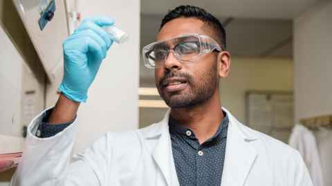 Man in a laboratory environment holding a container and wearing a white lab coat and safety glasses.