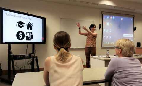 Liza in a lecture theatre gesturing towards a screen showing the words 'Socio-economic status'. Two people are in the foreground.