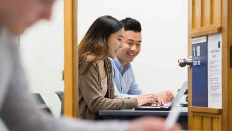 Two students seen through a doorway work together at a laptop