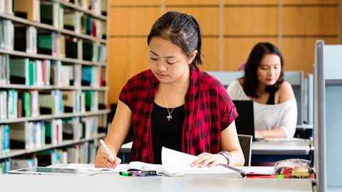 A female student sitting a desk highlighting text on paper in front of her. To the side is a book shelf and sitting behind her is another female working on a laptop