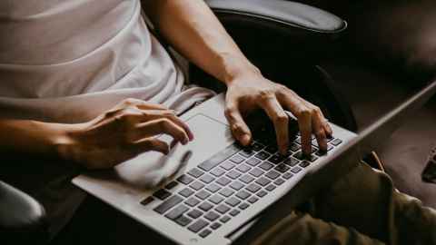 Lower arms and the lower half of a torso of a person wearing a white t-shirt. They are sat in a office chair with leather arms and have a laptop open on their lap