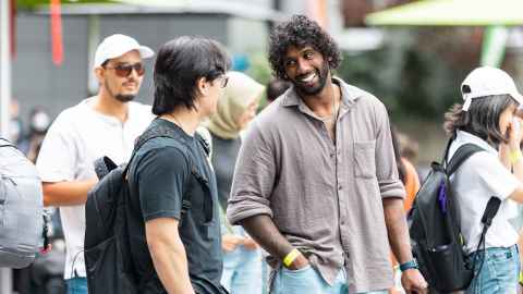 One student in black t-shirt carrying a black backpack talking to student wearing grey shirt at postgraduate orientation. Other students in the background.