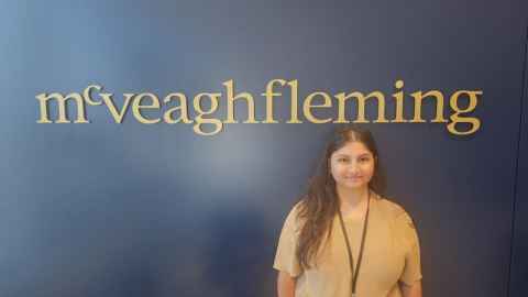 A woman with long dark hair wearing a beige tshirt standing in front of a dark blue background with a golden McVeagh Fleming sign