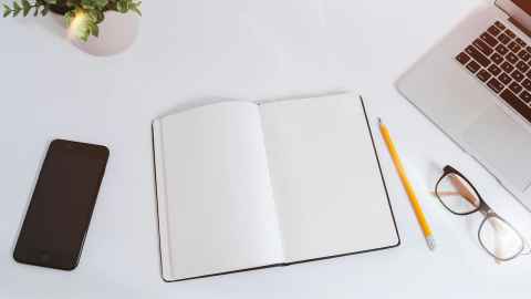 Desktop with various items on it - open notebook, pencil, phone, laptop and eyeglasses 