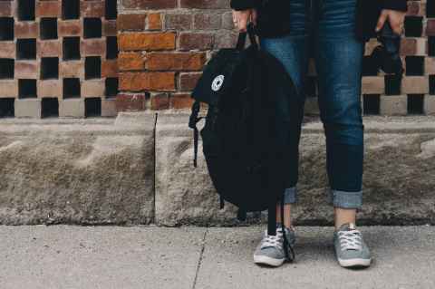 A photo from the hip down showing a person wearing jeans standing against a brick wall. They are holding a backpack