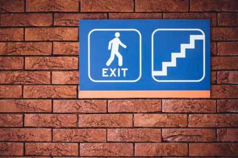 A brick wall with an exit sign split in two. One has a person walking, the other has stairs.