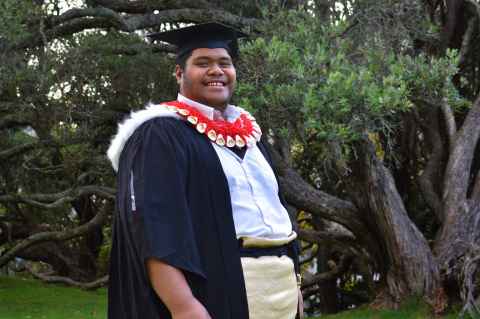 Soakai stands in front of a tree in his graduation regalia and traditional Tongan dress