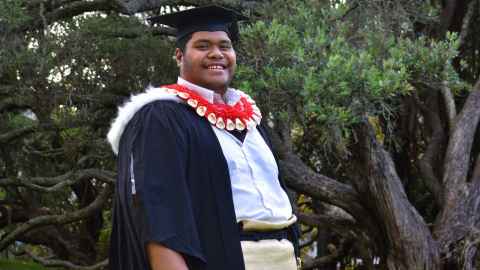 Soakai stands in front of a tree in his graduation regalia and traditional Tongan dress