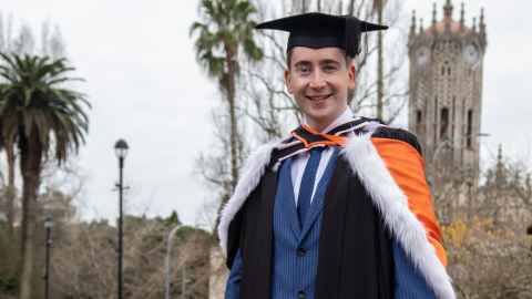 A young man wearing graduation regalia, including orange and pink hoods, with the ClockTower in the background.