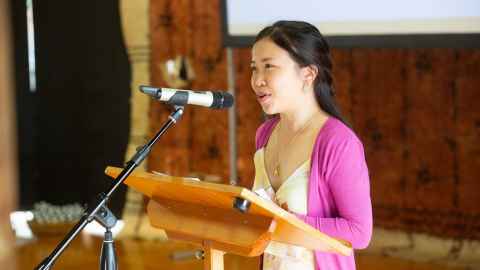 A young woman at a lectern, speaking into a microphone.