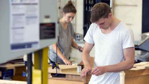 A young man, with a woman in the background, working with wood.