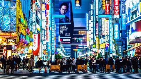 A crowded street at night, with electronic billboards and lighted signs, with writing in an Asian language.