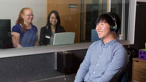 A young man in a booth with headphones on, smiling, and two women looking on, also smiling.