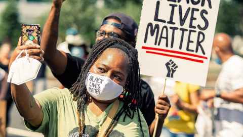 A woman in a mask taking part in a Black Lives Matter protest holding a phone before her.