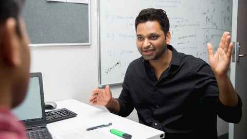 A man sitting at a desk with a laptop, talking to another man.