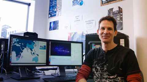 A smiling man sitting at a desk with computer screens.