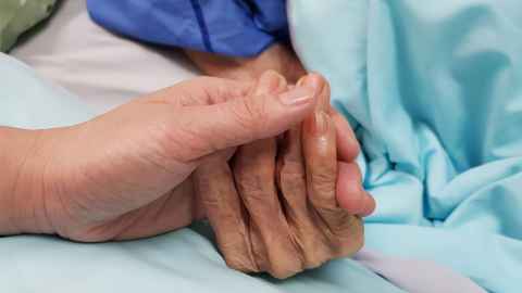 A person holding the hand of an elderly patient in a hospital bed.