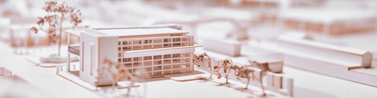 A close-up view of an architectural scale model featuring a modern, rectangular building with large grid-like windows and flat roofs. The model includes miniature trees and surrounding structures, all in a monochromatic, light-toned color scheme, giving a clean and minimalistic appearance.