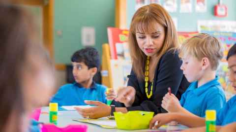 A women sitting at a table in a classroom with several children.