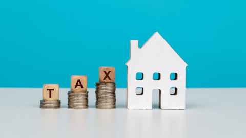 A one-dimensional wooden model of a house next to piles of coins with the letters T, A and X on wooden blocks sitting on top of them.