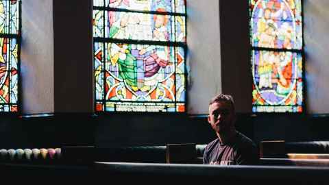 A young man sitting in a church with stained glass windows.