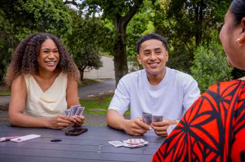 Three Pacific people sitting at an outdoor table, playing cards.