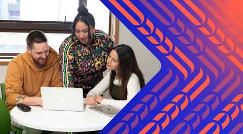 Two women and a man sitting around a table looking at a laptop screen together