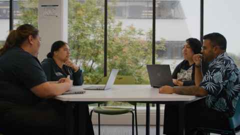 A group of three women and one man sitting around a table talking to each other with laptops open in front of them