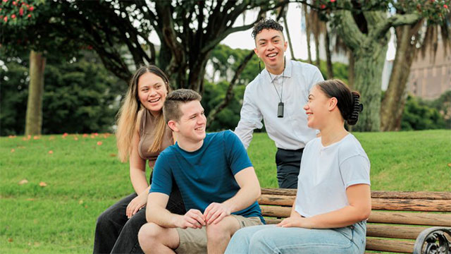 Four students talking, with two seated on a park bench and two standing