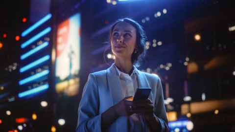 Beautiful Young Woman Using Smartphone Standing on the Night City Street Full of Neon Light. Portrait of Gorgeous Smiling Female Using Mobile Phone.